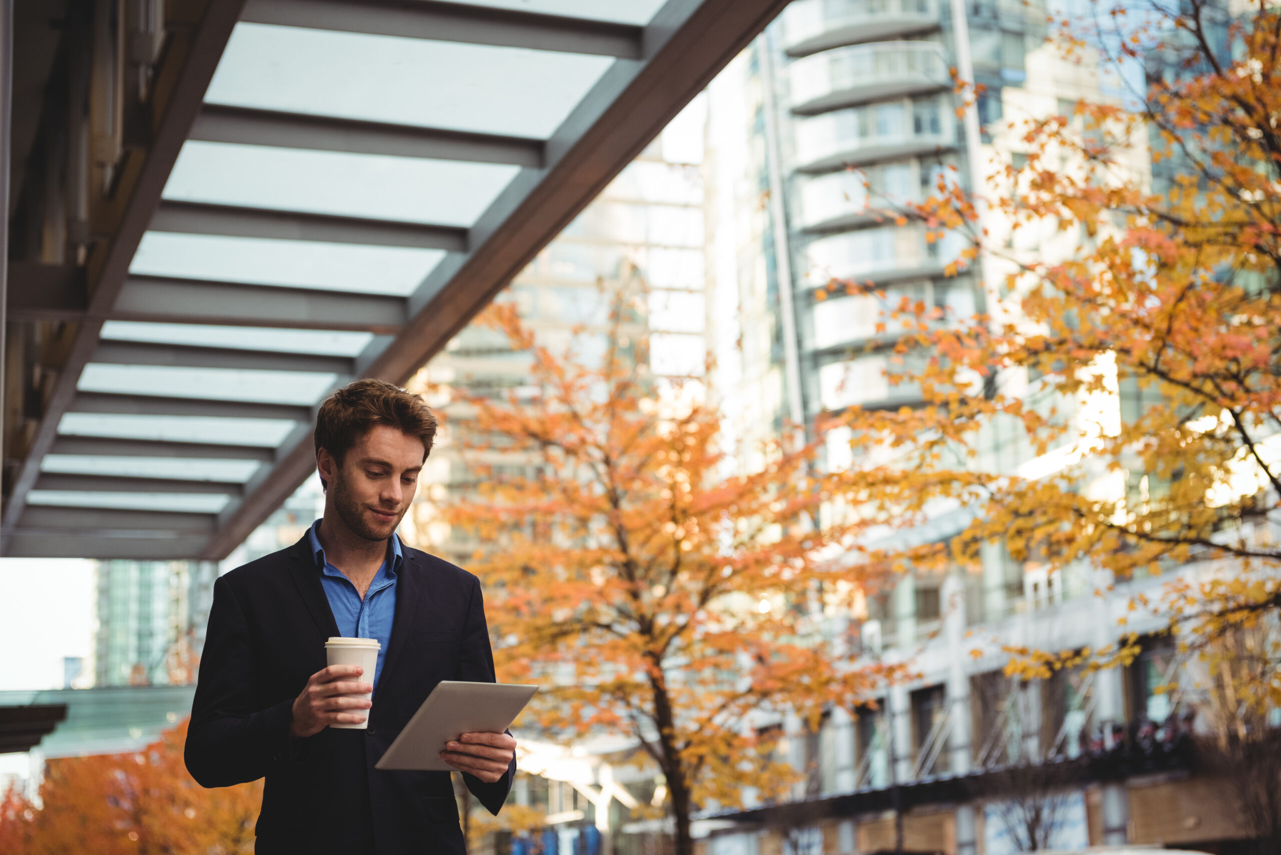 Businessman holding disposable coffee cup and using digital tablet while walking on the street